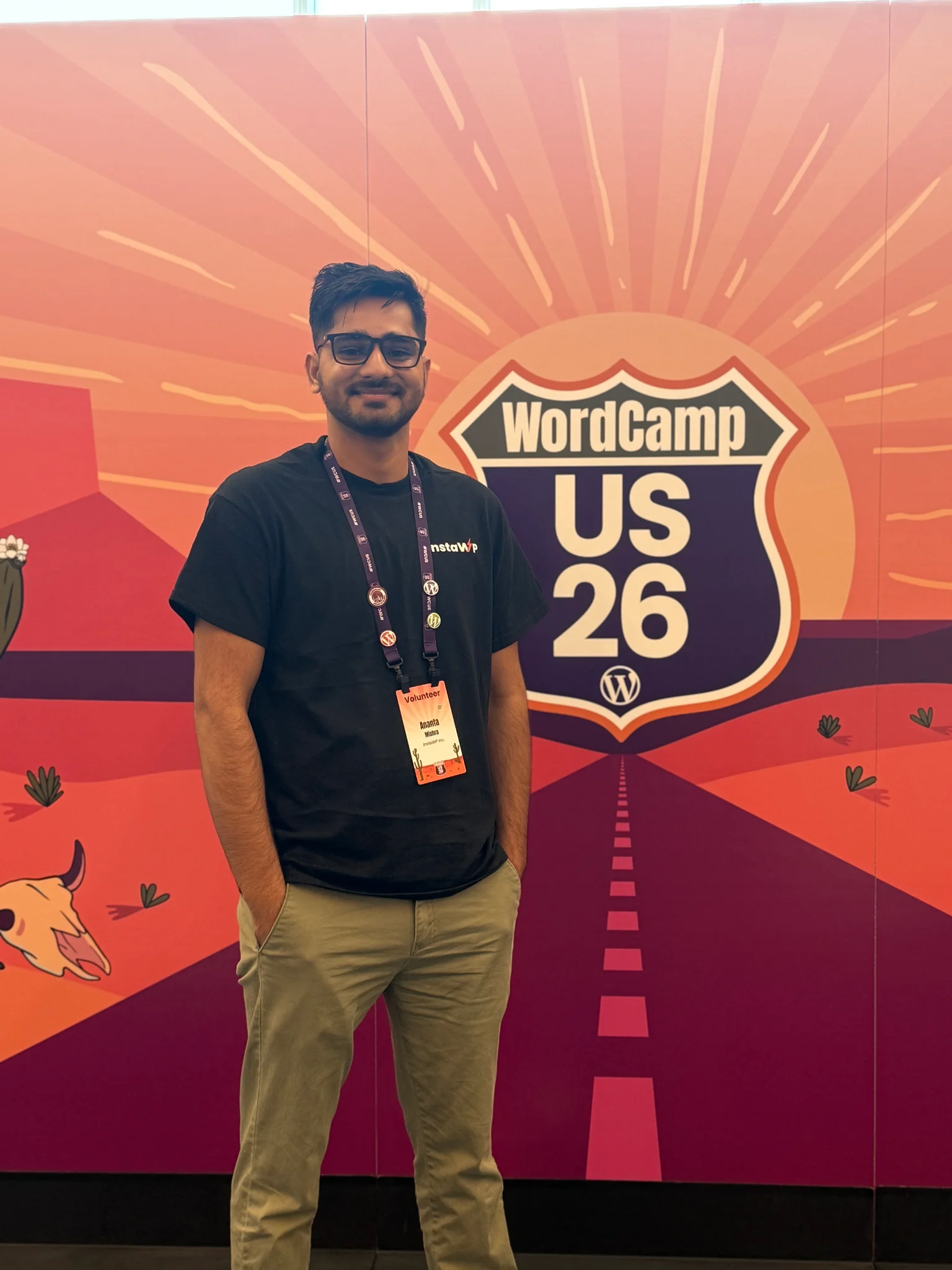 Ananta smiling in front of the WordCamp US 26 desert sunset highway banner